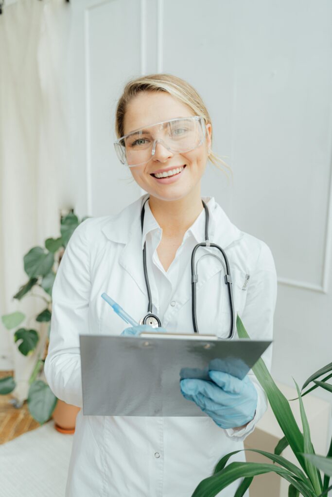 Smiling healthcare professional wearing safety glasses, a stethoscope, and gloves, holding a clipboard in a clinic setting.